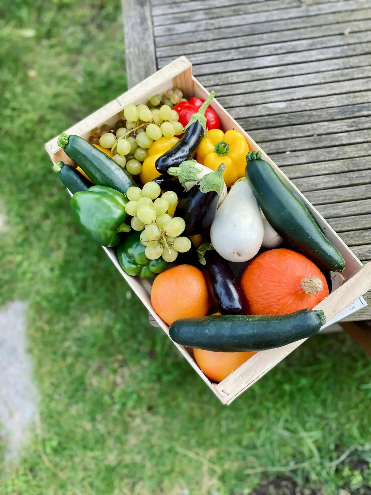 A wooden crate of squash, peppers, grapes and eggplant on the grass, seen from above