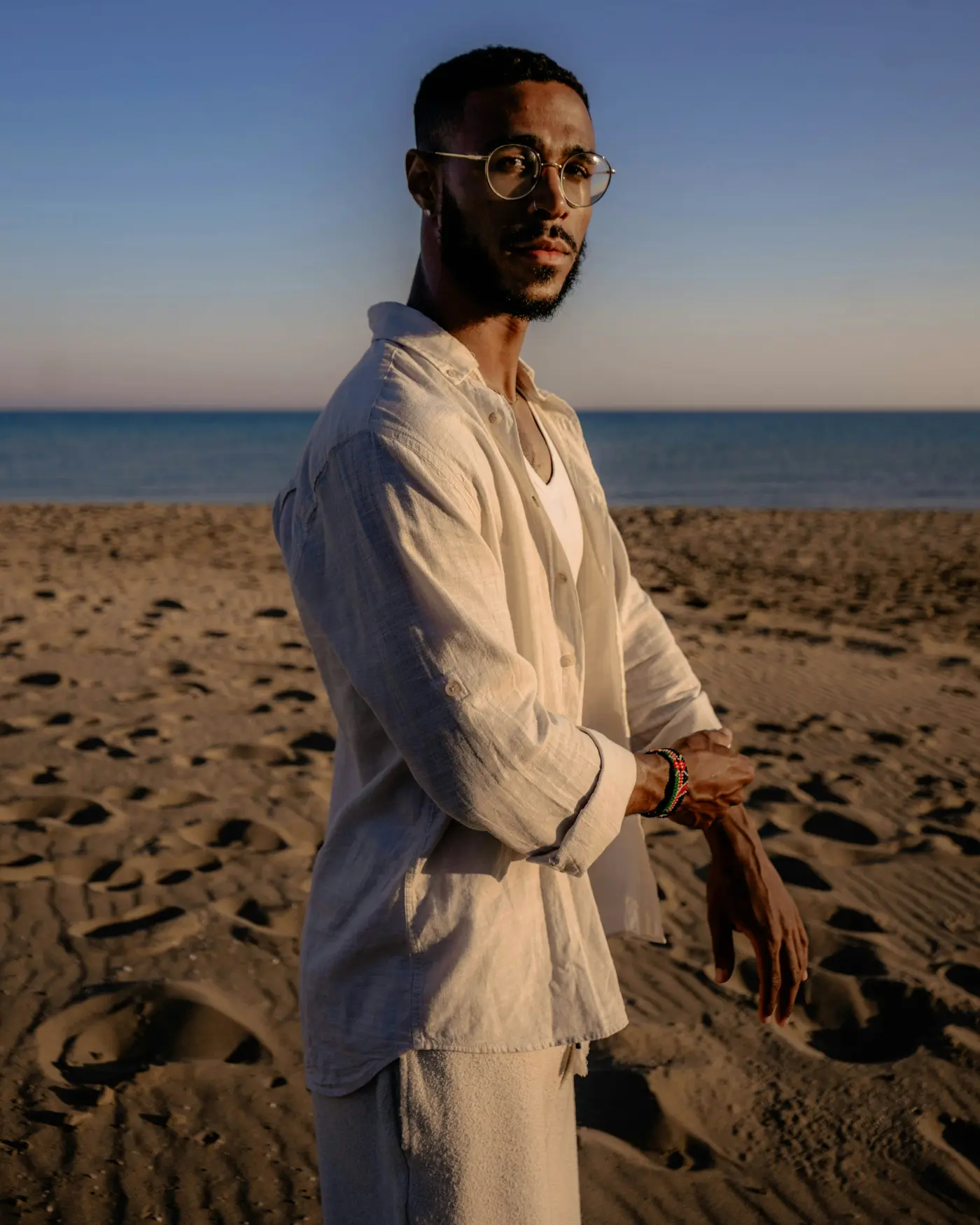 A man in a white linen shirt and trousers on a beach at sunset