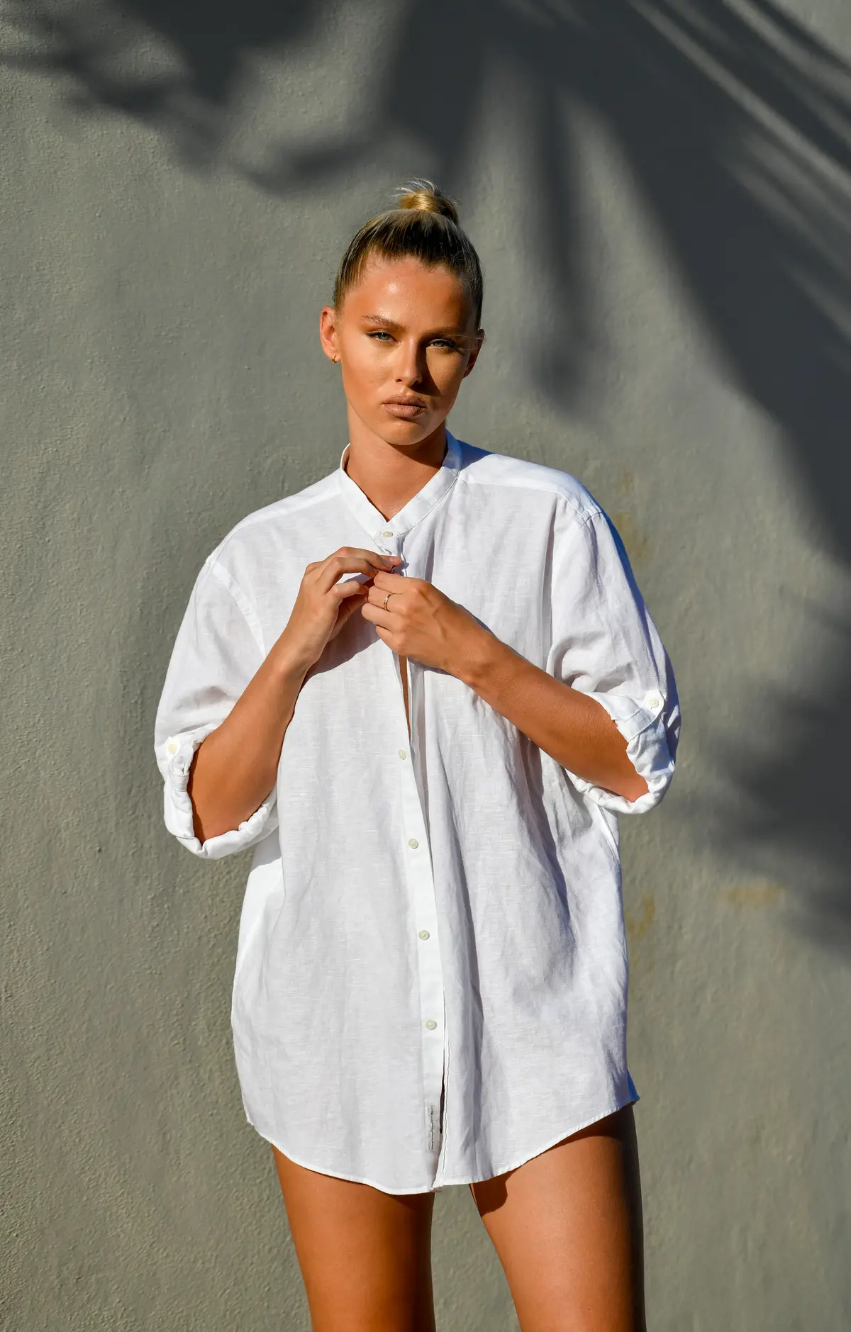 A woman in an oversized white linen shirt against a sunlit wall