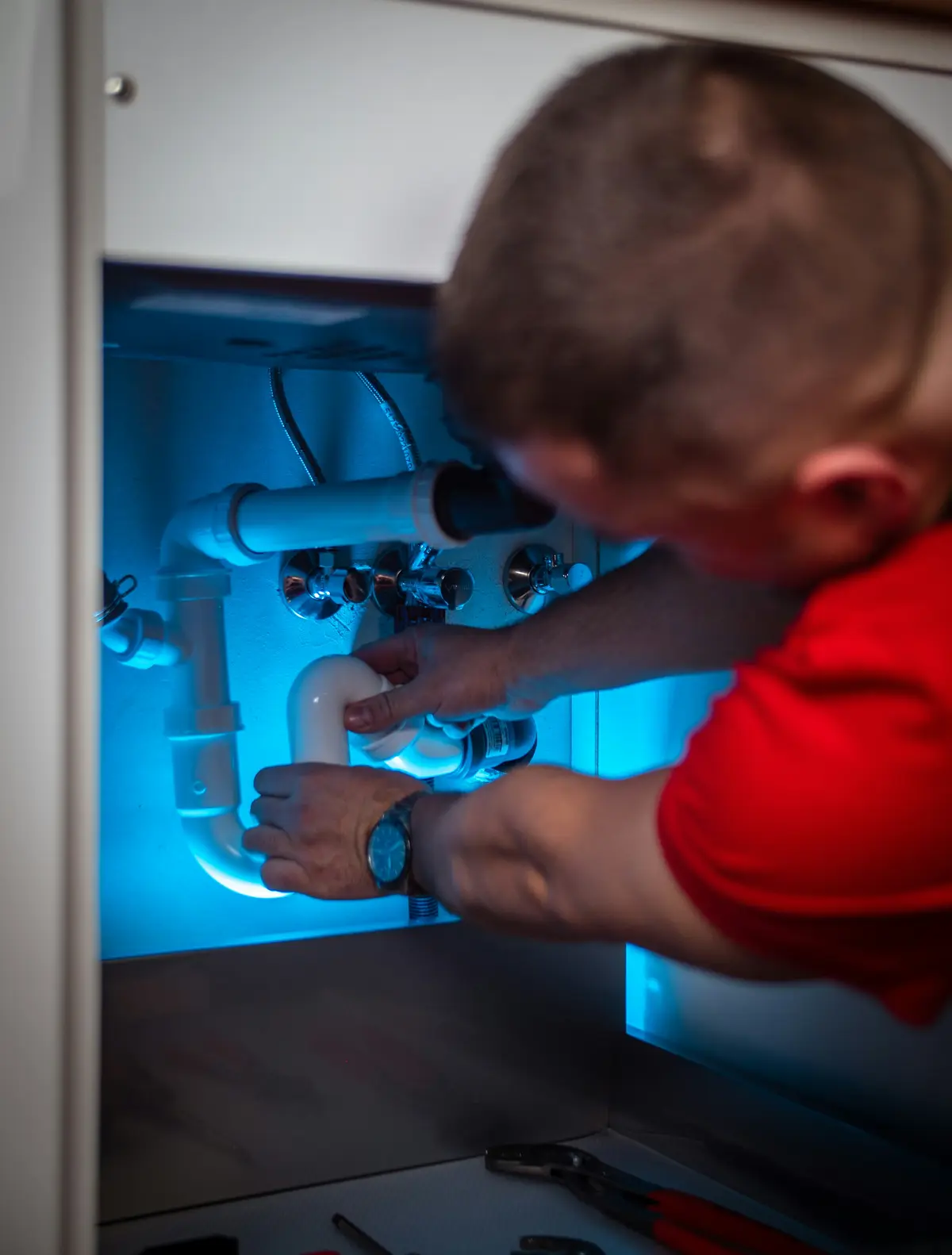 A plumber in a red shirt reaching into a sink cabinet lit blue