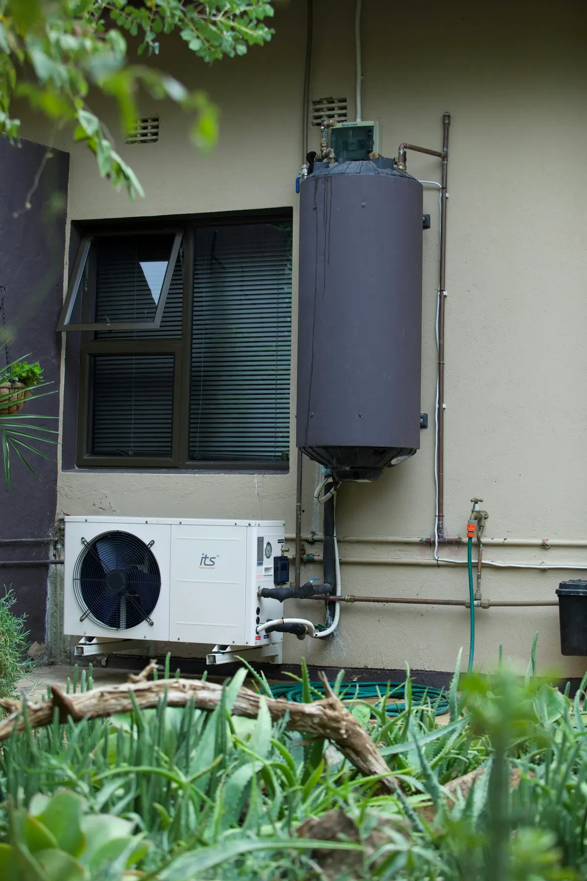 An outdoor unit and a tank water heater on the side of a house, behind green plants