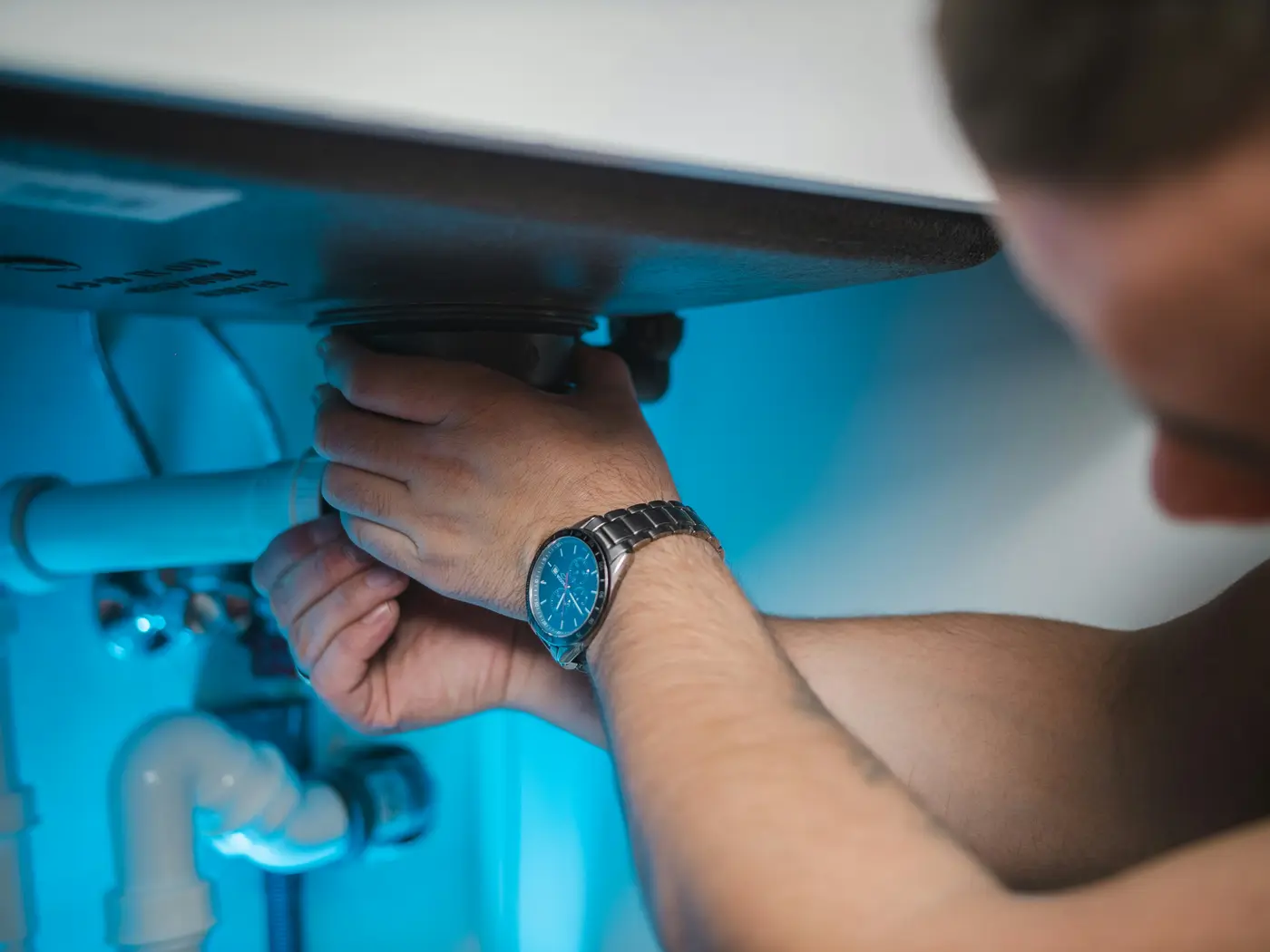 A plumber’s hands tightening a fitting under a sink, lit blue