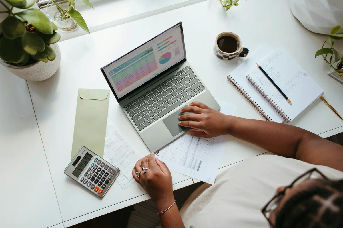 Hands at a laptop next to a calculator, a notebook and a coffee, seen from above