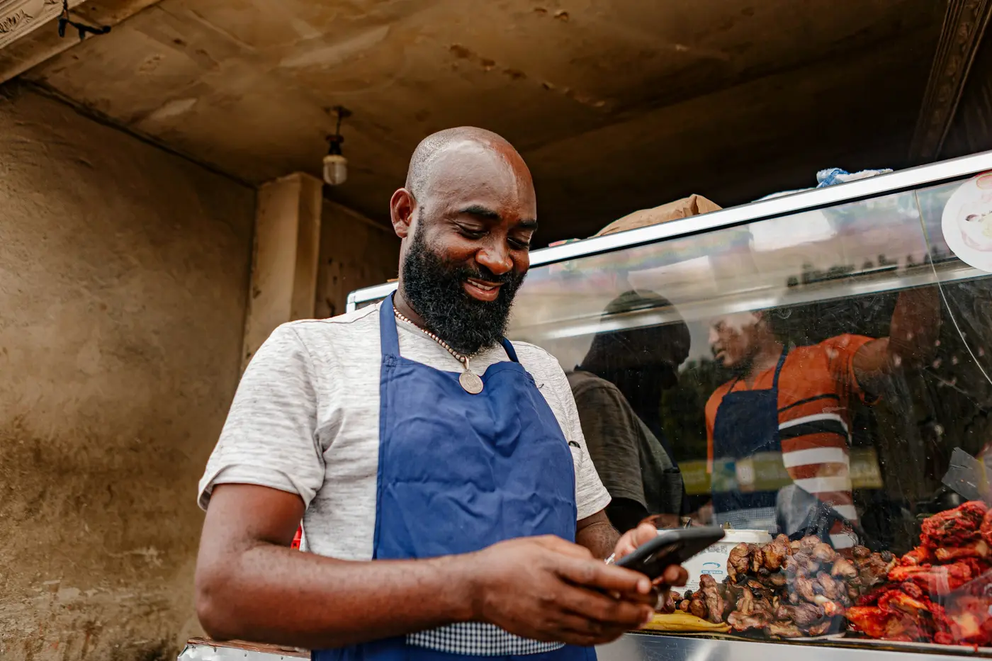 A butcher in a blue apron smiling at his phone behind the meat counter