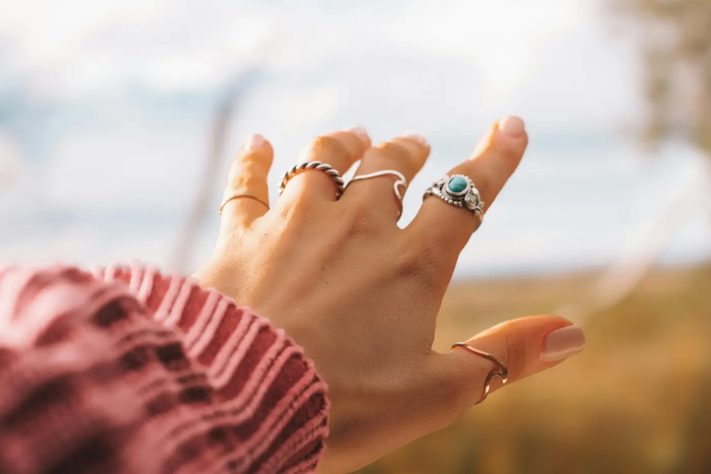 A hand wearing several thin rings, reaching toward a field at sunset