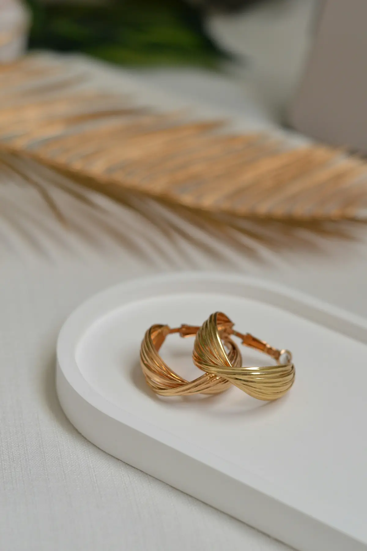 Twisted gold hoop earrings on a small white tray beside a dried leaf