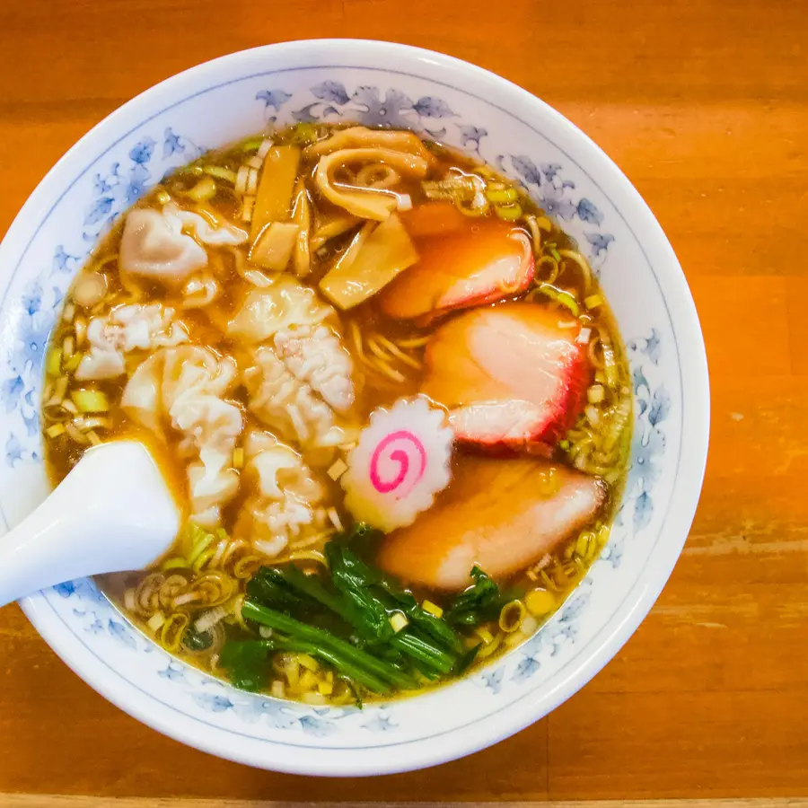 A clear shoyu ramen with pork, fish cake and greens in a patterned bowl