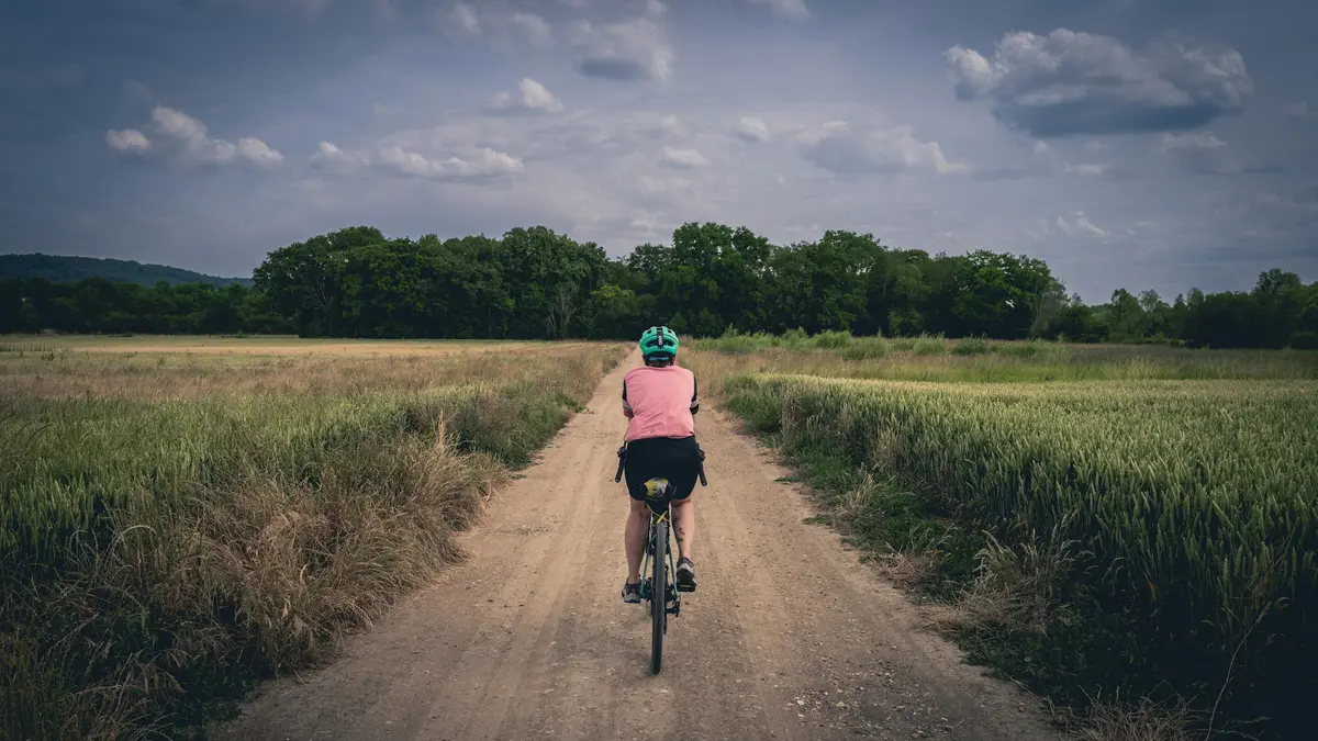 A rider on a long straight dirt road between fields under a grey sky